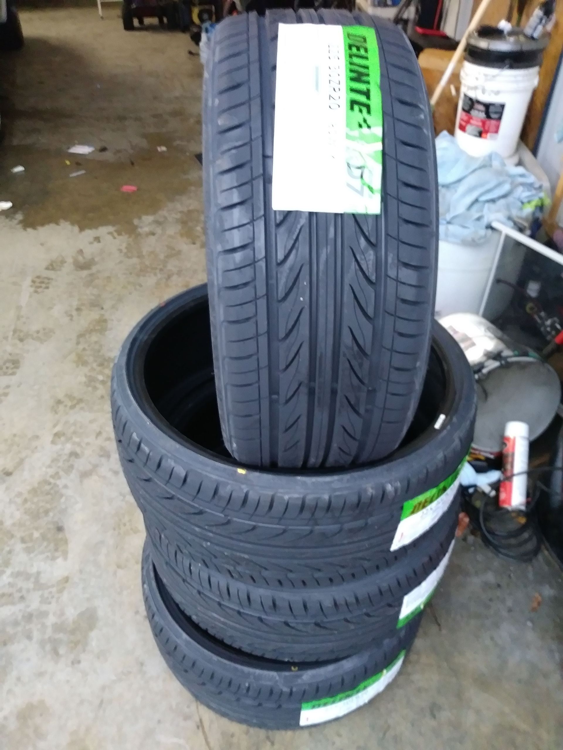 Four new black tires stacked on top of each other, in a garage setting. Each has a green and white label.