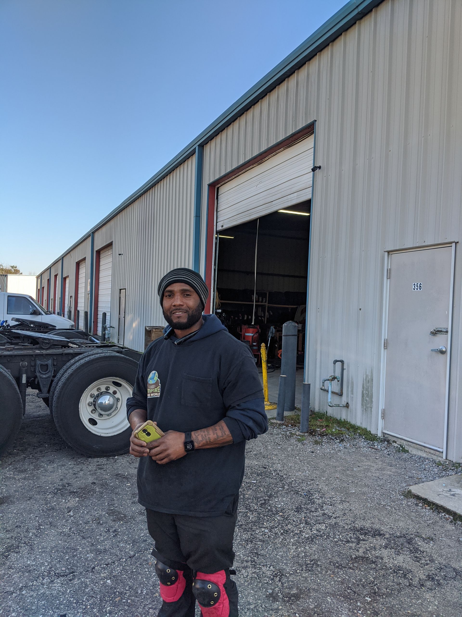 Man in front of a repair shop holding a yellow object, with a semi truck and open garage door in the background.