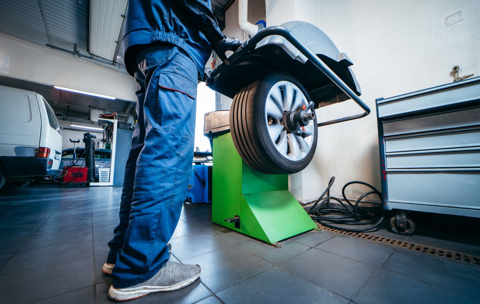 Mechanic balances a car tire on a green machine in a garage; blue work clothes.
