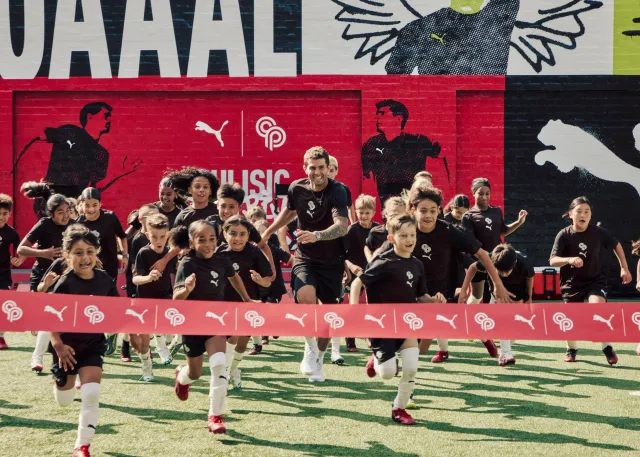 Soccer team posing on the field in front of a red mural, with players running toward the camera in celebration