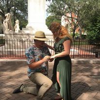 A man is kneeling down to propose to a woman in front of a statue.