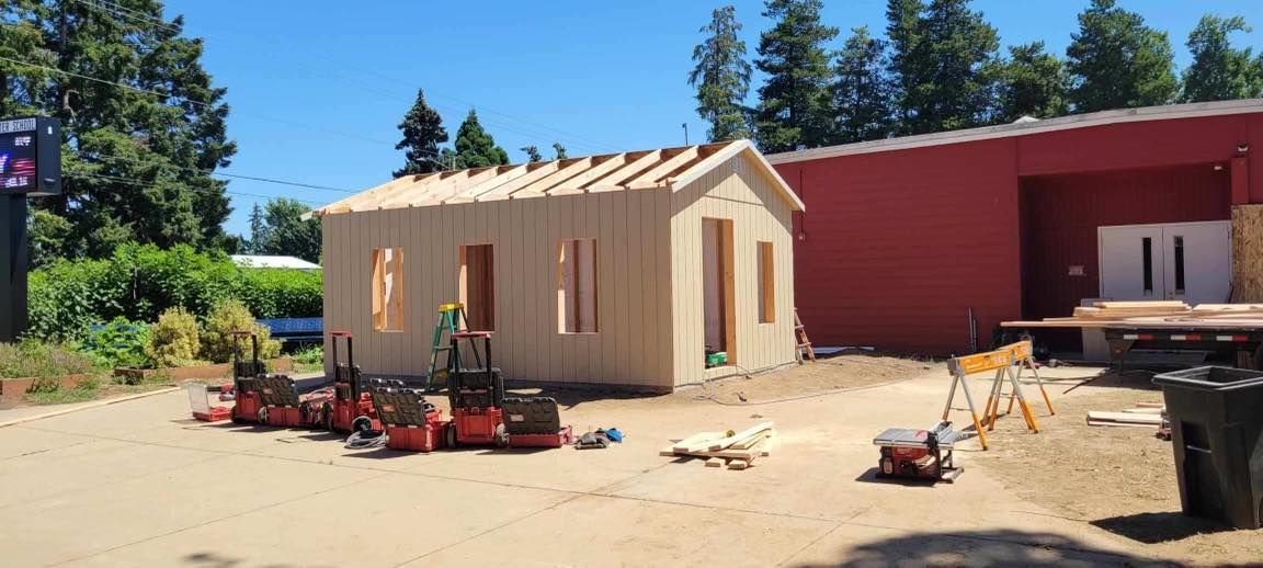 A small wooden house is being built in a dirt lot in front of a red building.