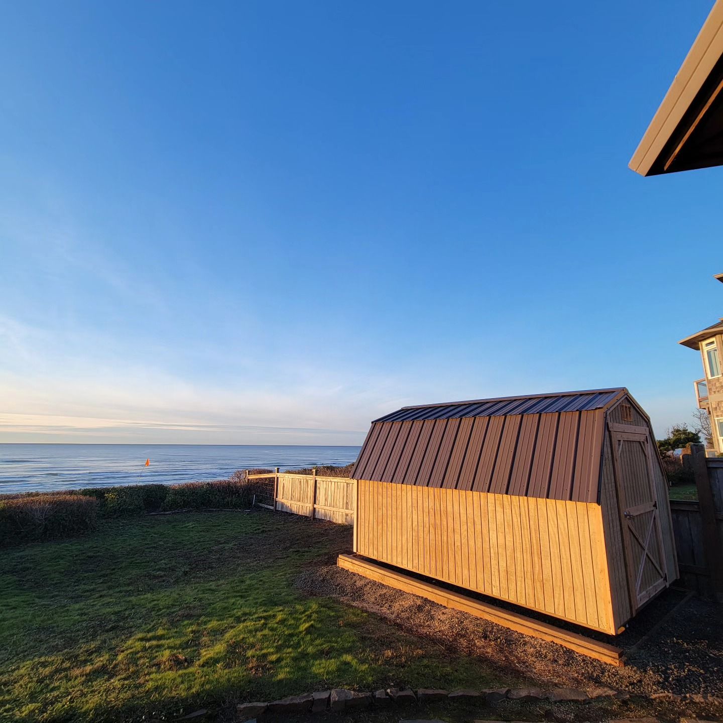 A wooden shed is sitting in the grass next to a house with a view of the ocean.
