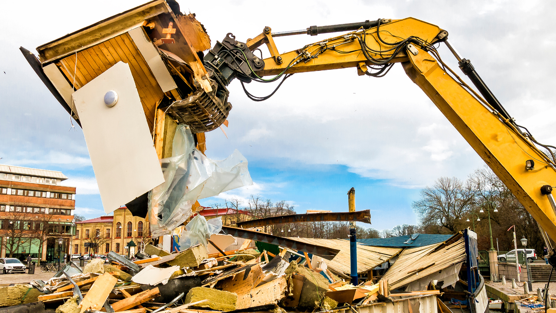 A bright yellow construction excavator lifts a piece of building debris from a demolition site under a cloudy sky.
