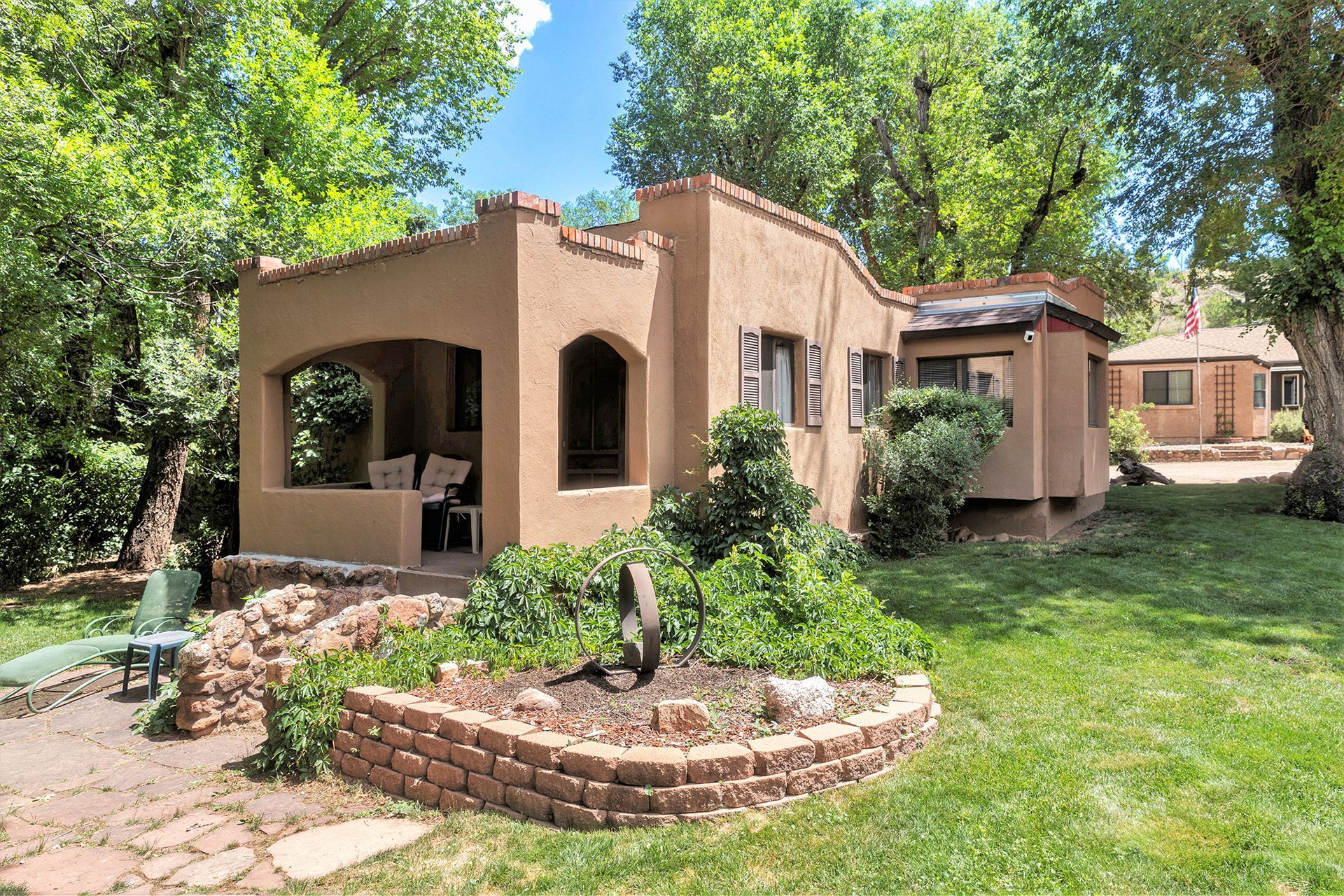 A small adobe house with a brick planter in front of it is surrounded by trees.