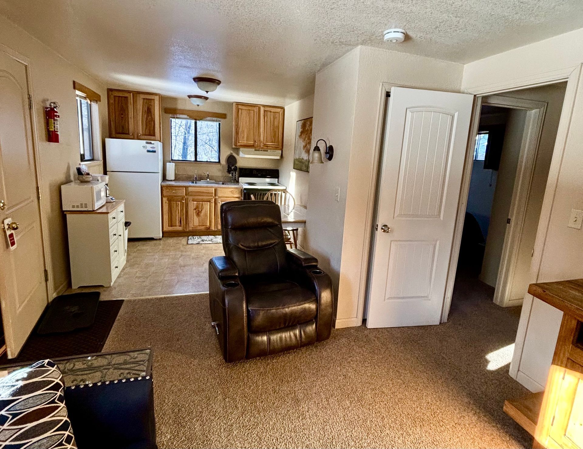 Cozy cabin interior: Kitchen with appliances, a brown recliner, a doorway, and beige carpet.