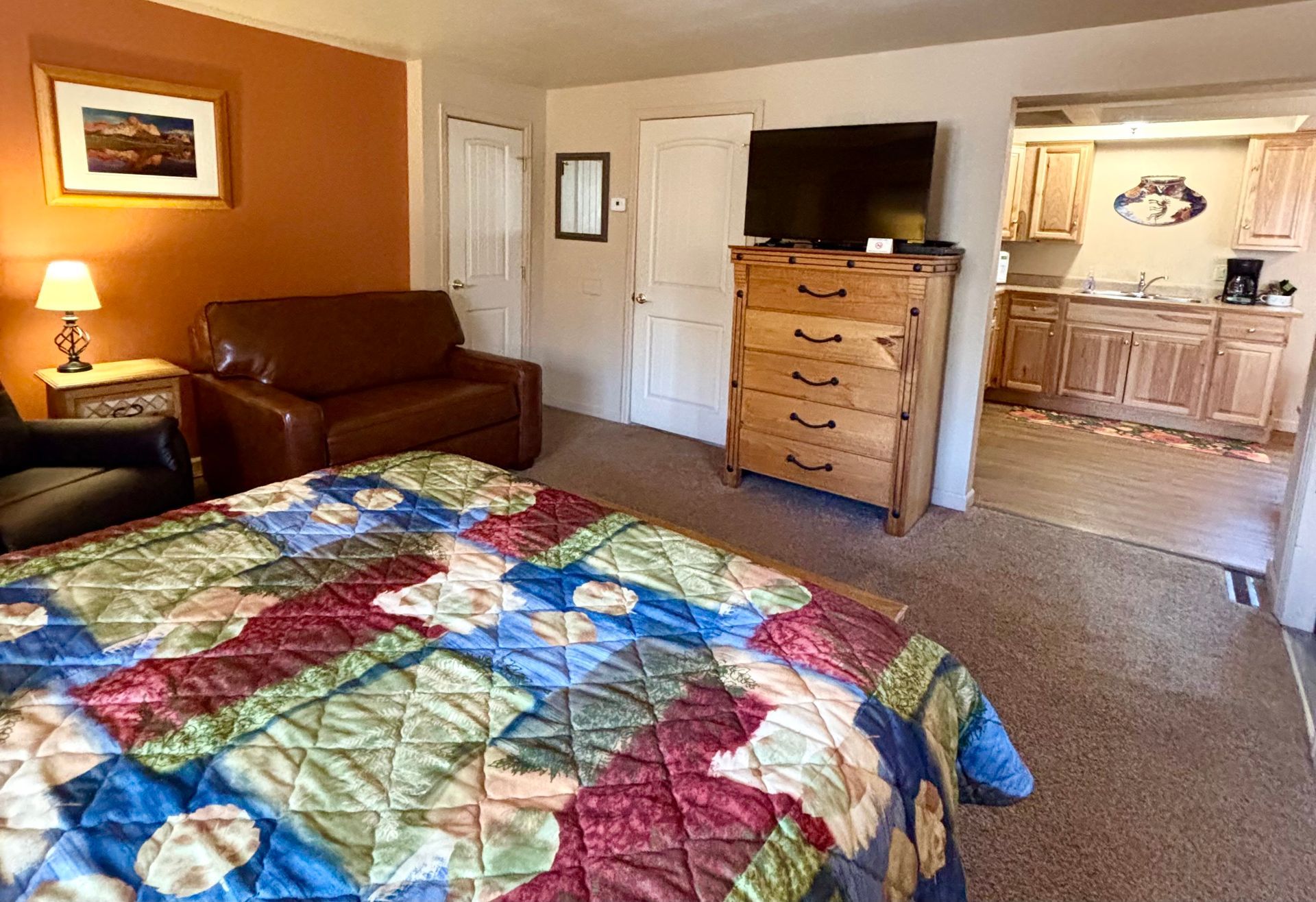 Hotel room with bed, sofa, dresser, TV, and kitchenette visible in background.