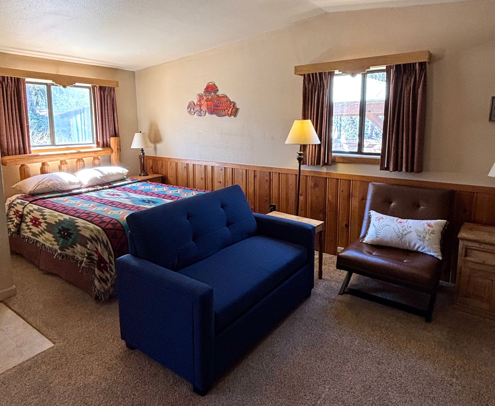 Bedroom with a bed, blue sofa, brown armchair, and two windows with brown curtains.
