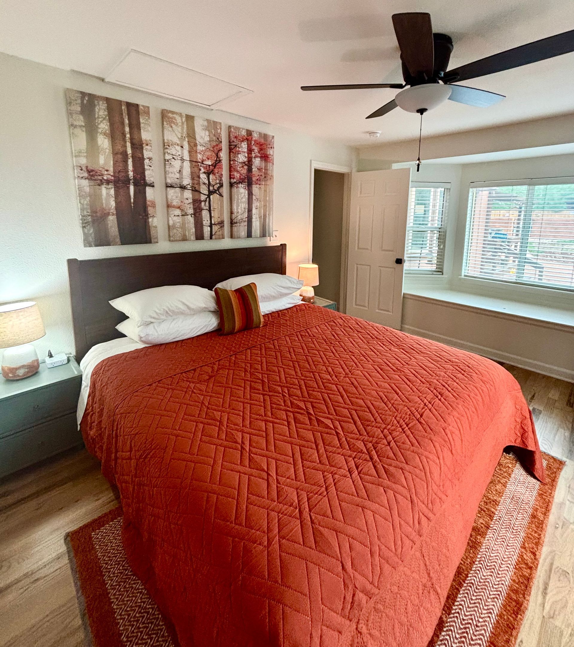 Bedroom with orange quilt, artwork, a window seat, and ceiling fan.