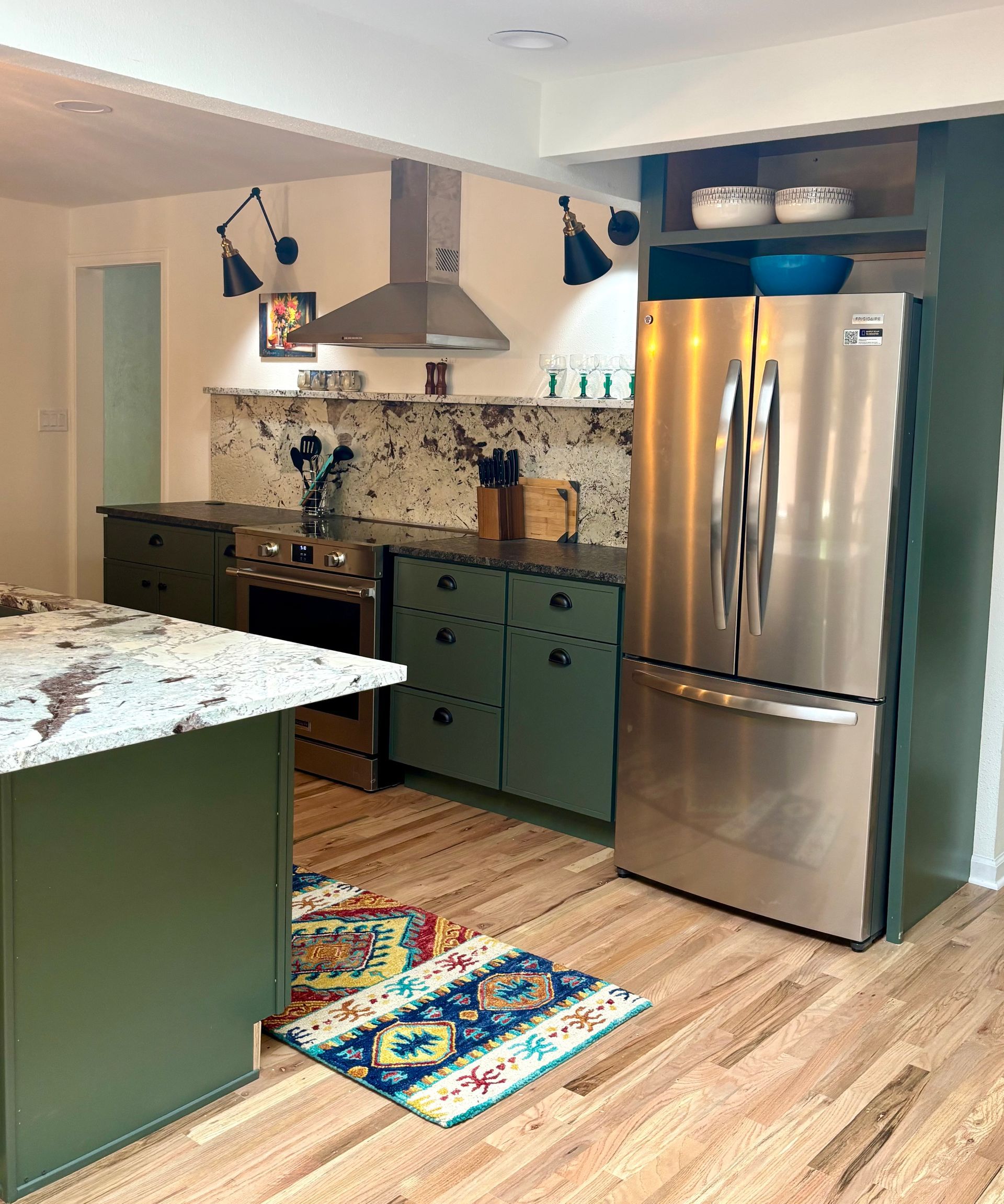 Green kitchen with stainless steel fridge, patterned backsplash, olive cabinets, and wood flooring.