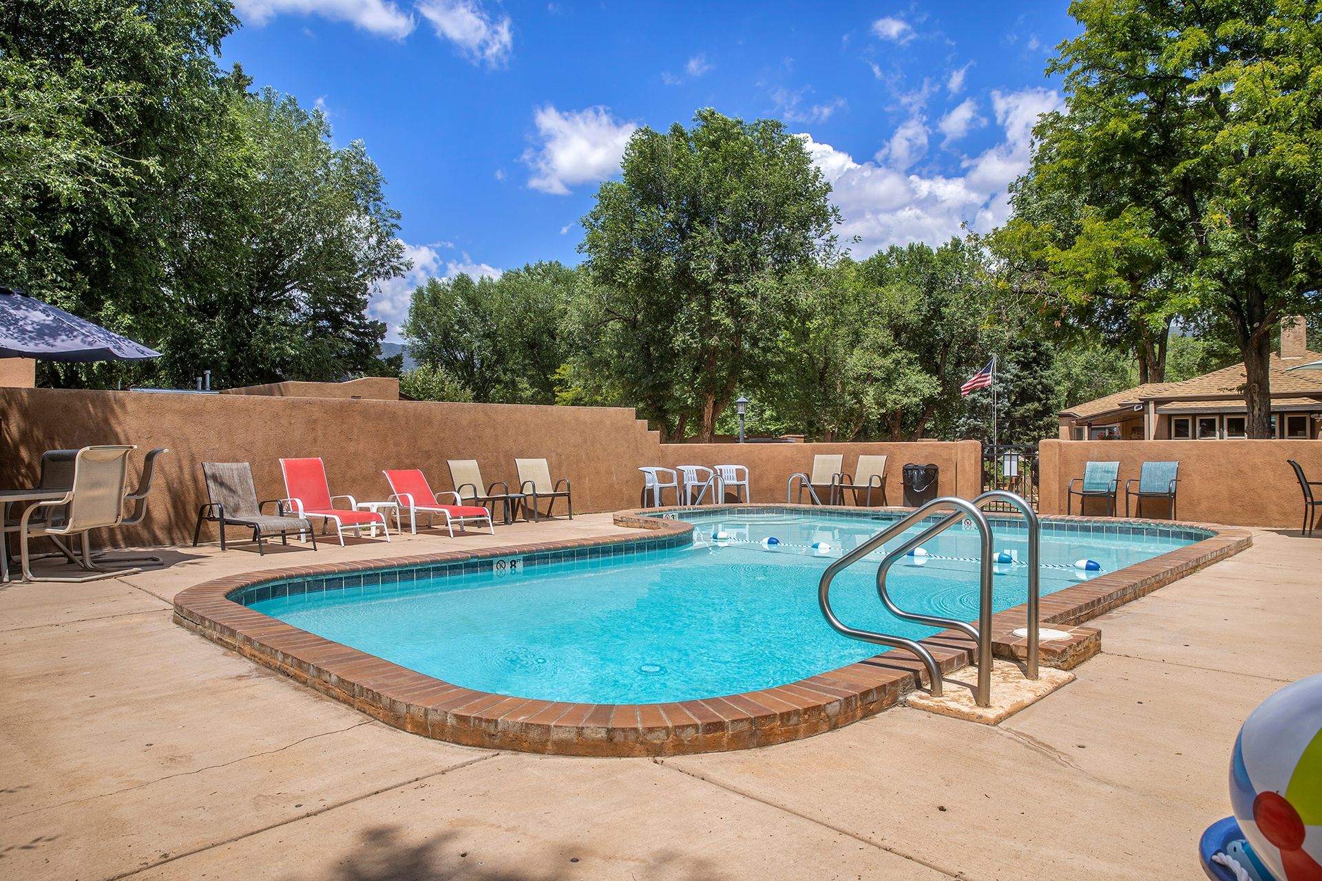 Swimming pool surrounded by chairs on a sunny day. A low wall and trees are in the background.