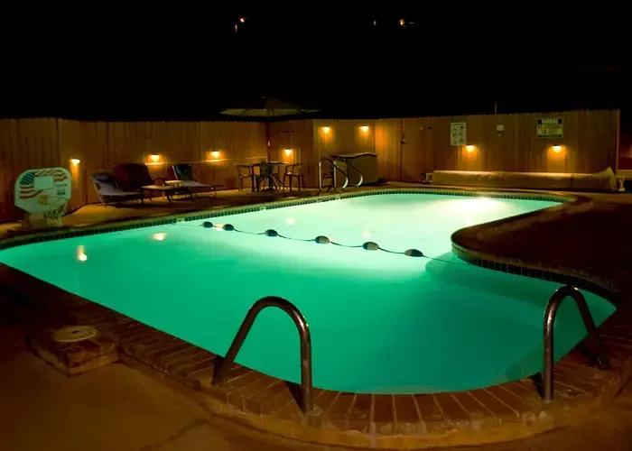 Nighttime view of a lit swimming pool surrounded by a wooden fence, lights, and lounge chairs.