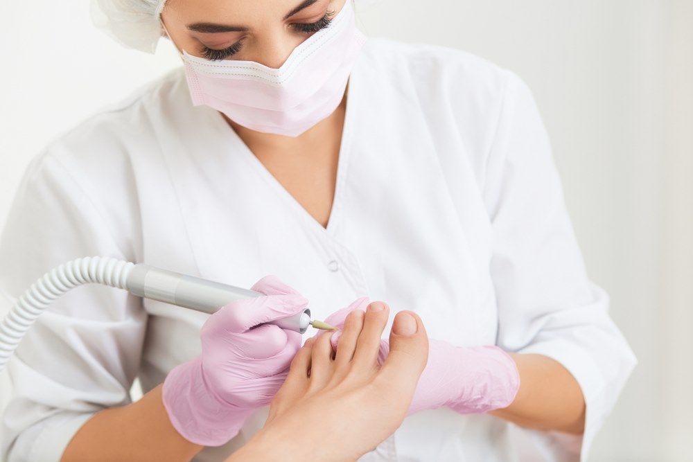 A Woman is Wearing a Mask and Gloves While Doing a Foot Treatment — Maitland Central Podiatry in Maitland NSW