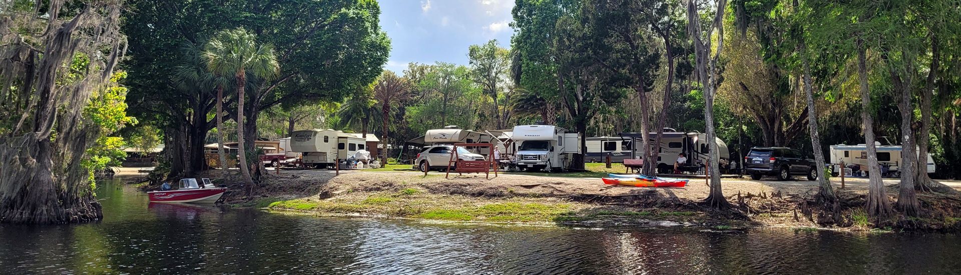a row of rvs are parked on the shore of a lake surrounded by trees .