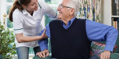 A nurse is helping an elderly man sit on a couch.