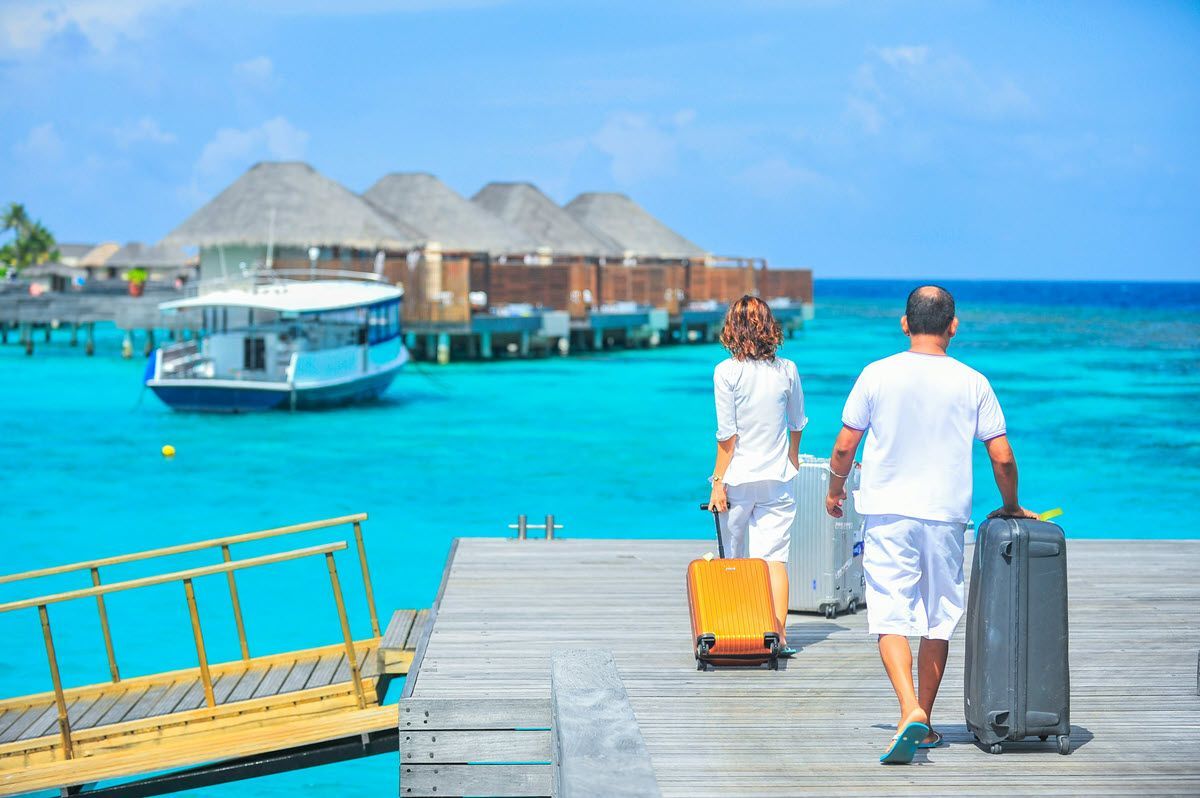 Couple walking on a wooden dock with luggage toward overwater bungalows and a boat in bright turquoise water
