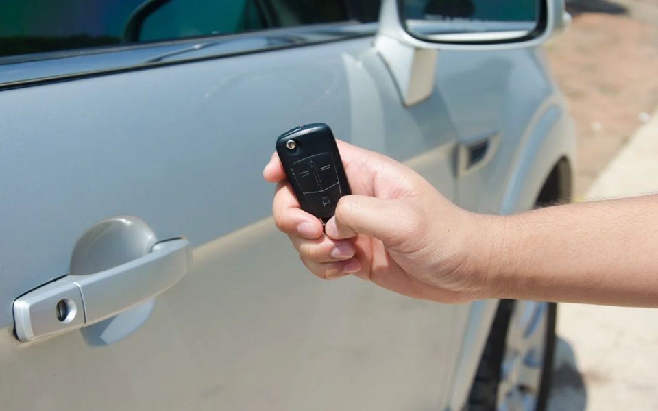 A hand holding a smart key in front of a silver car.