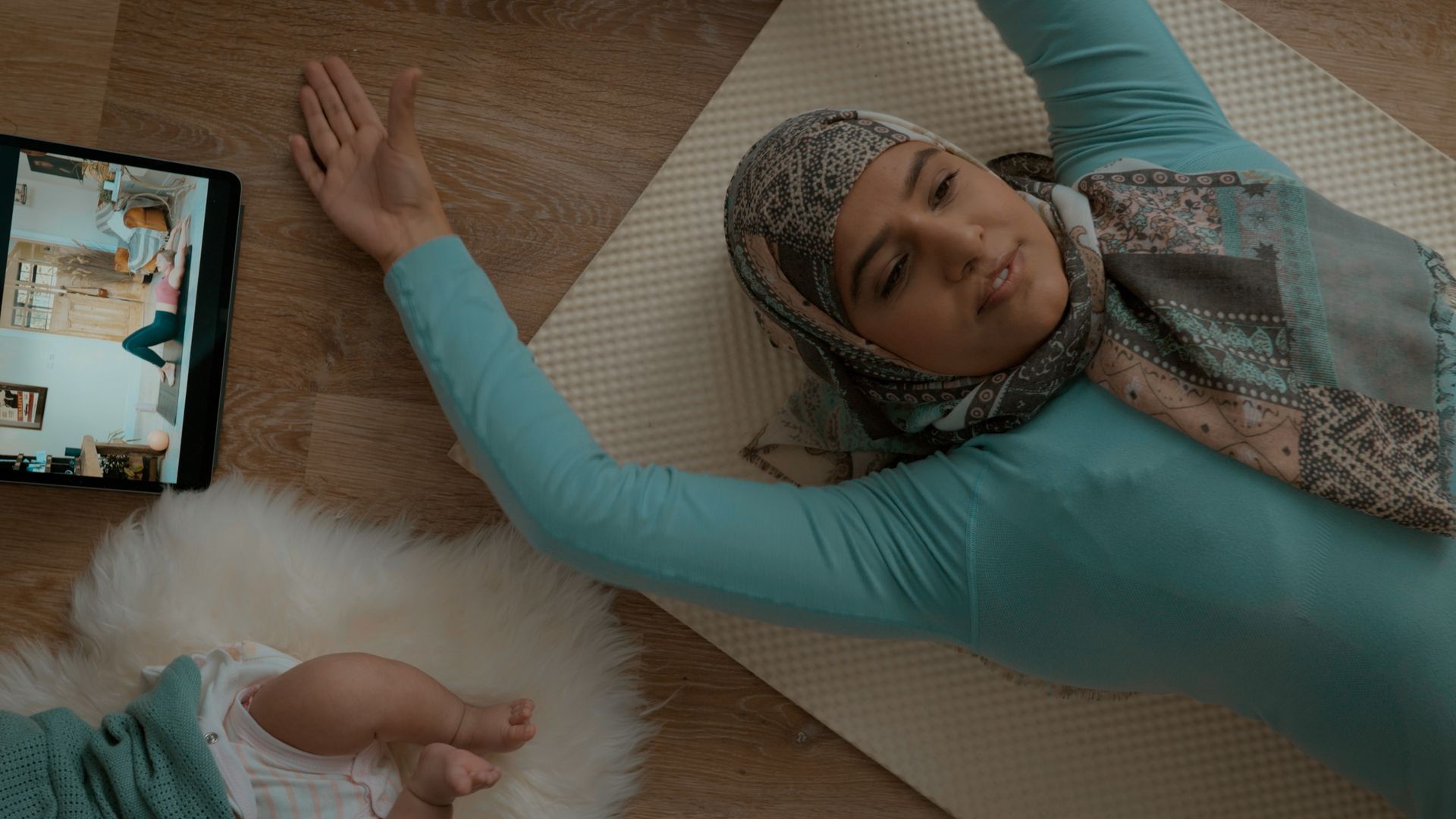 A woman in a hijab is laying on a yoga mat next to a baby.