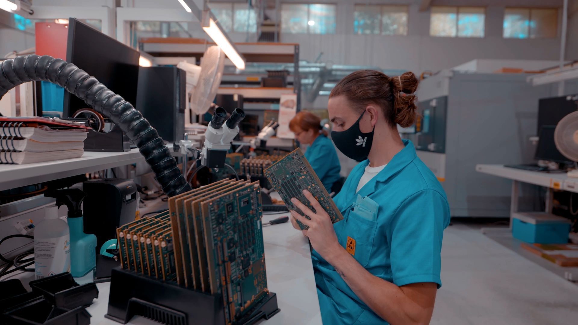 A woman wearing a mask is holding a motherboard in a factory.