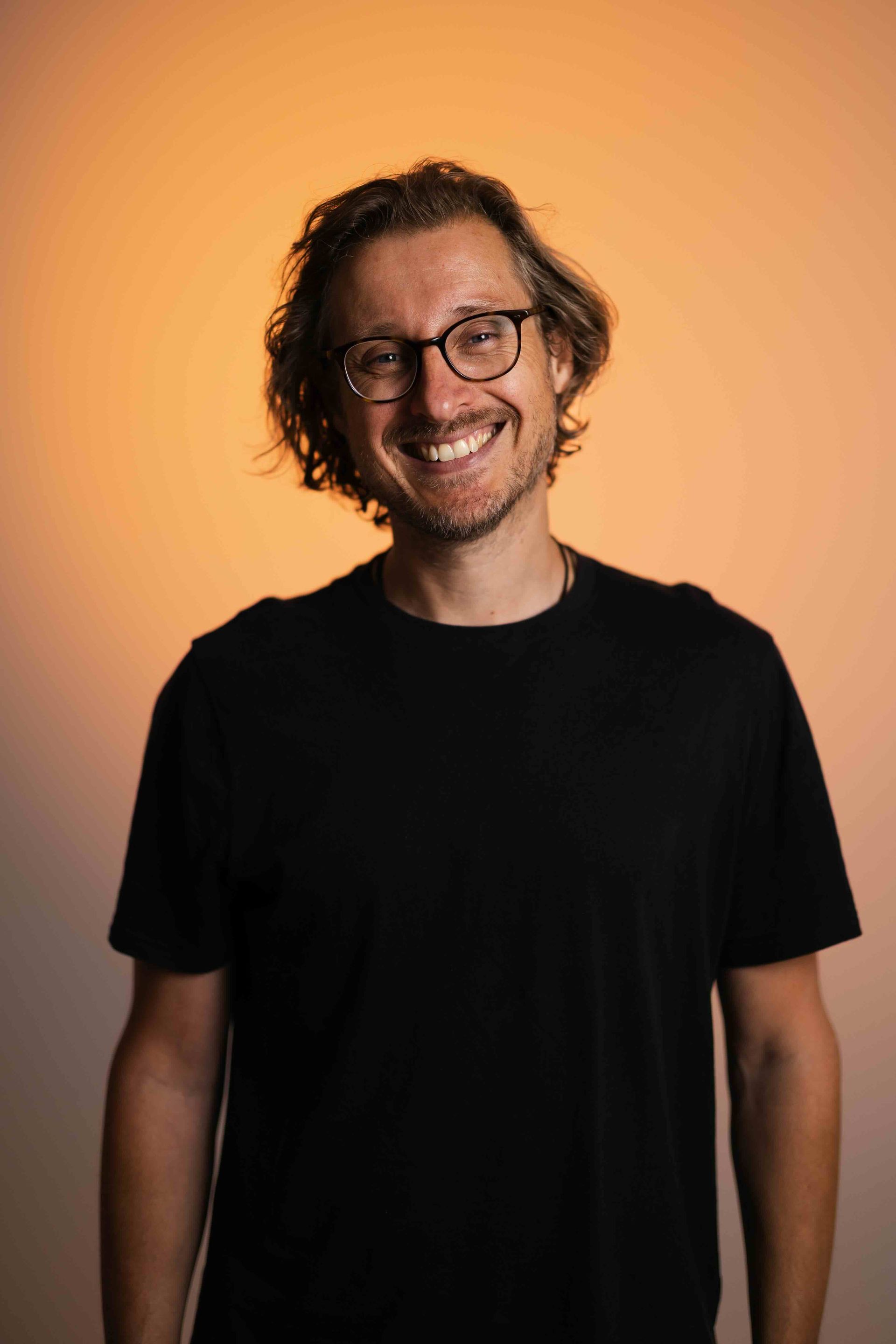 A man with long hair and a beard is wearing a gray shirt and smiling.