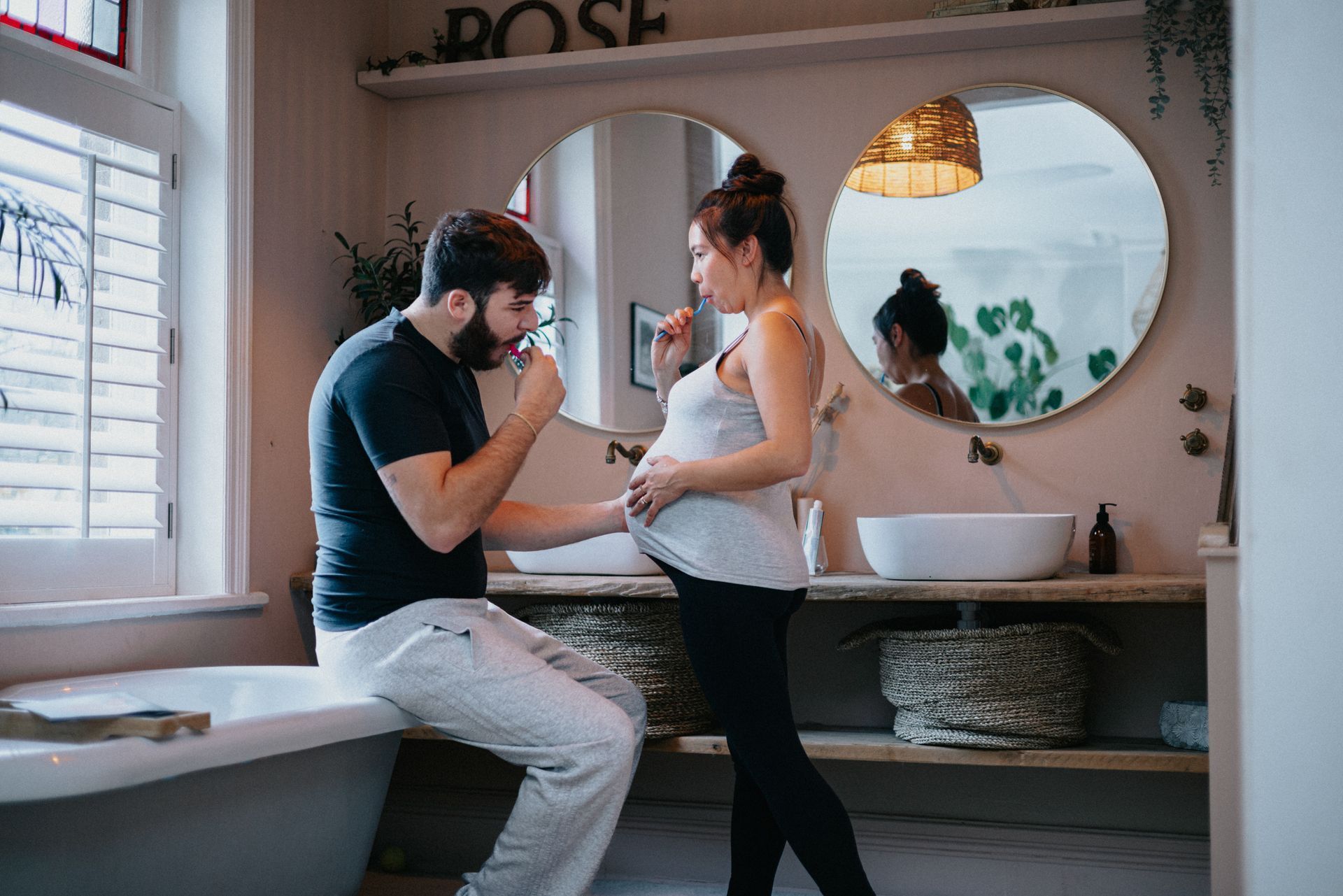 A man is brushing his teeth next to a pregnant woman in a bathroom.
