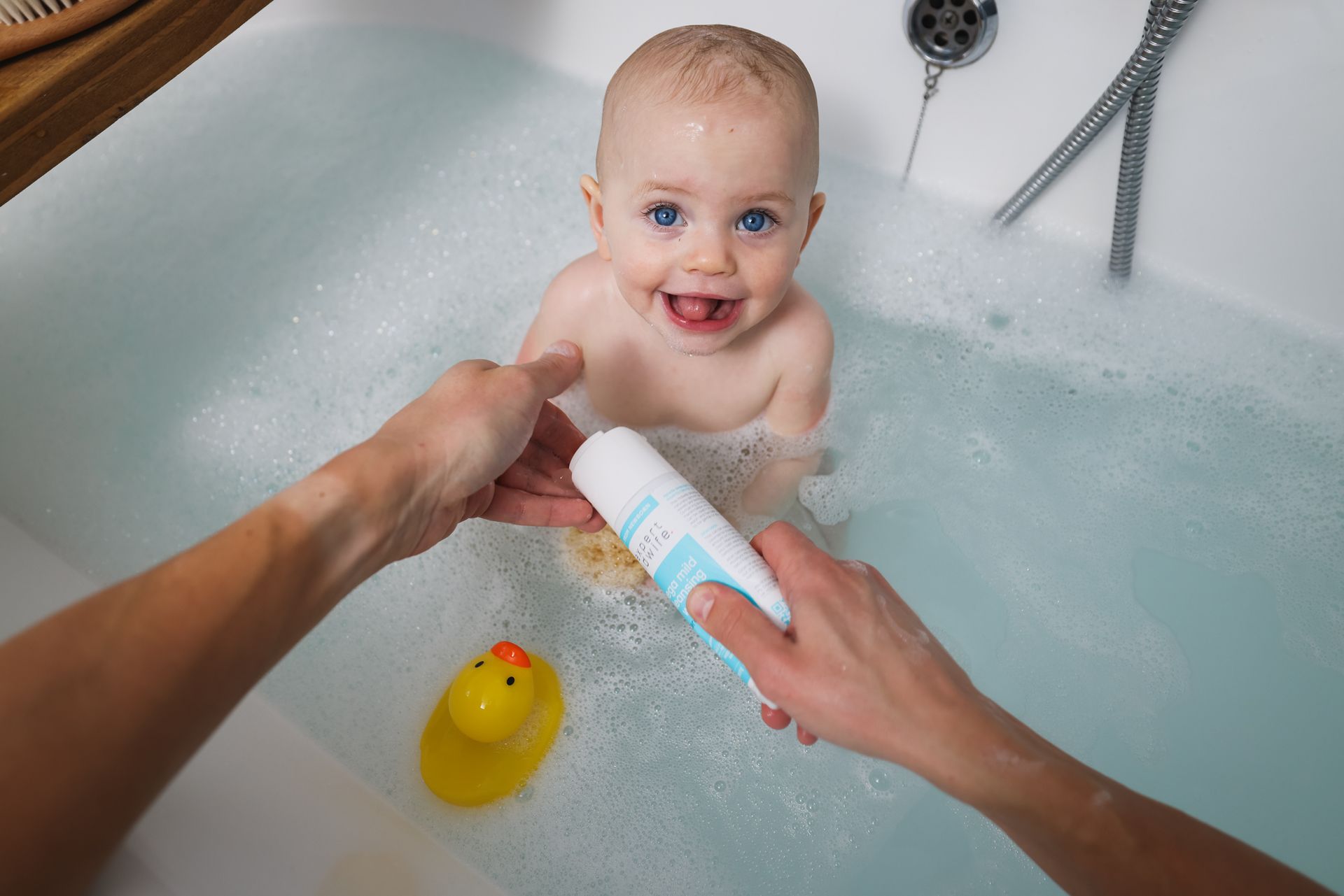 a baby is taking a bath in a bathtub while a person holds a bottle of shampoo .