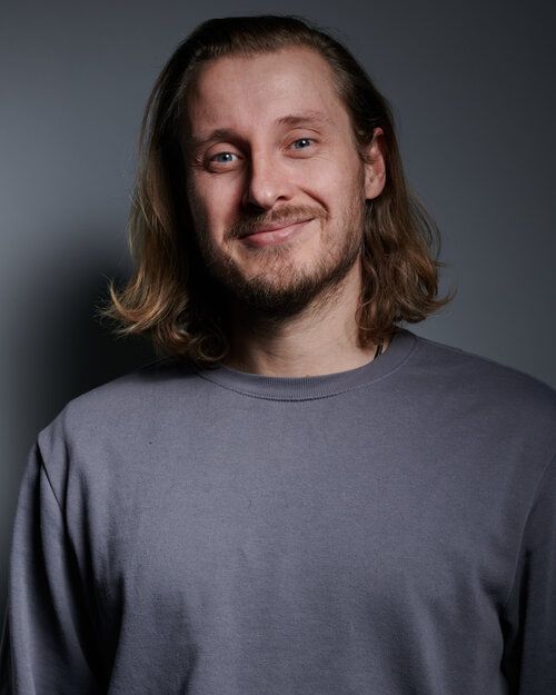 A man with long hair and a beard is wearing a gray shirt and smiling.