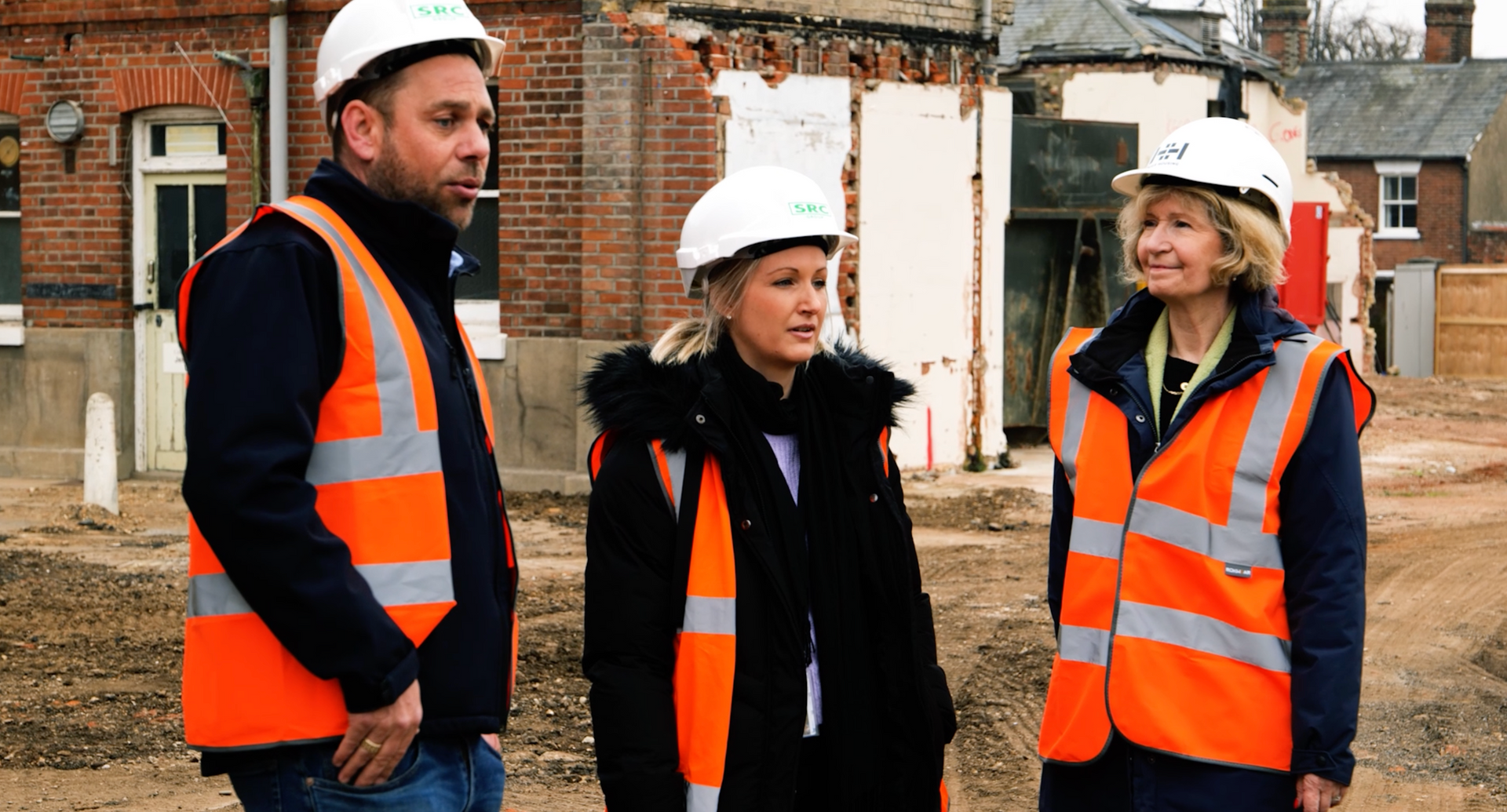 A man and two women wearing hard hats and orange vests are standing on a construction site.