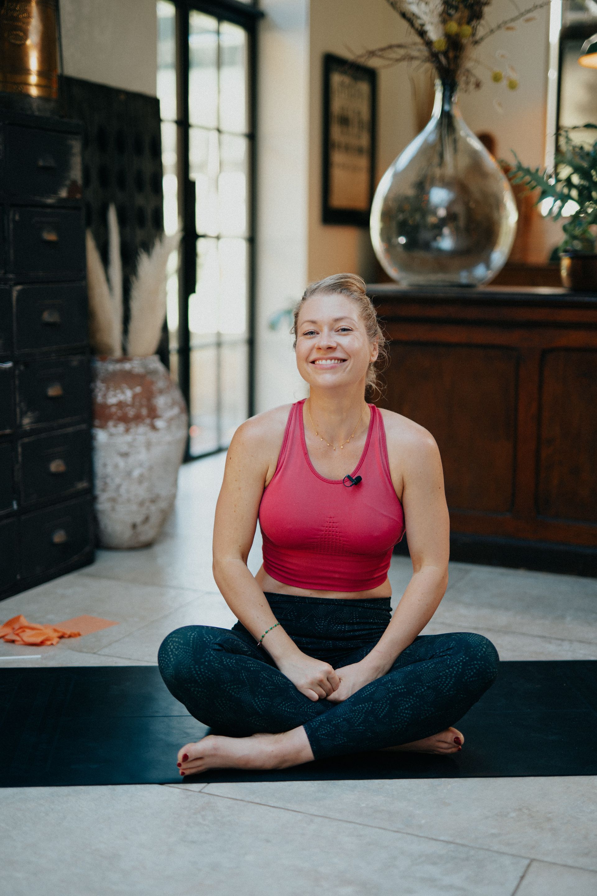 A woman is sitting on a yoga mat with her legs crossed and smiling.