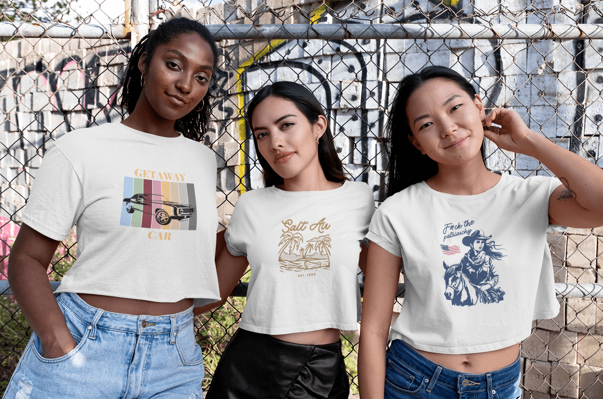 Three women wearing crop tops are standing next to each other in front of a chain link fence.