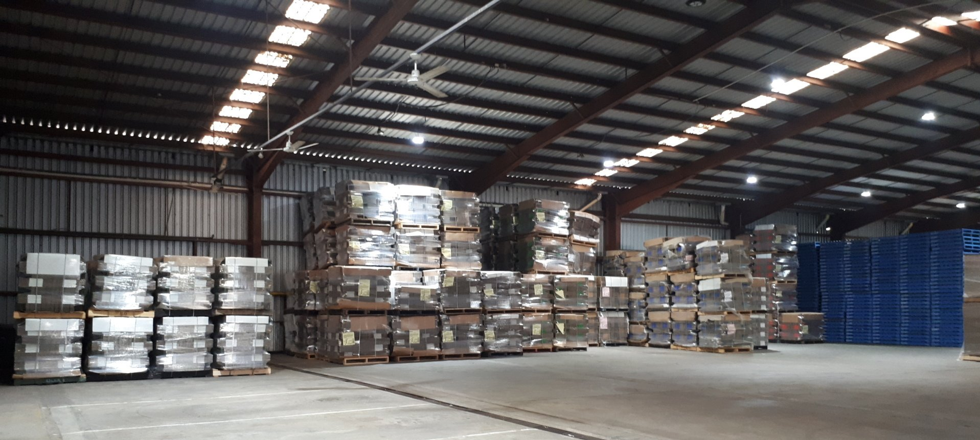 Warehouse interior with stacked goods; corrugated metal roof, packed pallets of supplies.