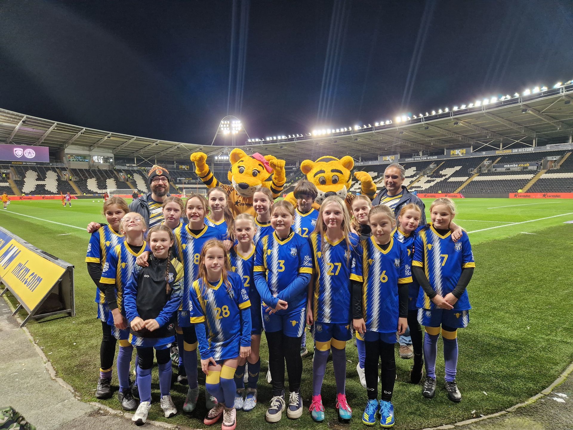 Youth sports team posing with mascots on a stadium field at night. Team wears blue and gold.