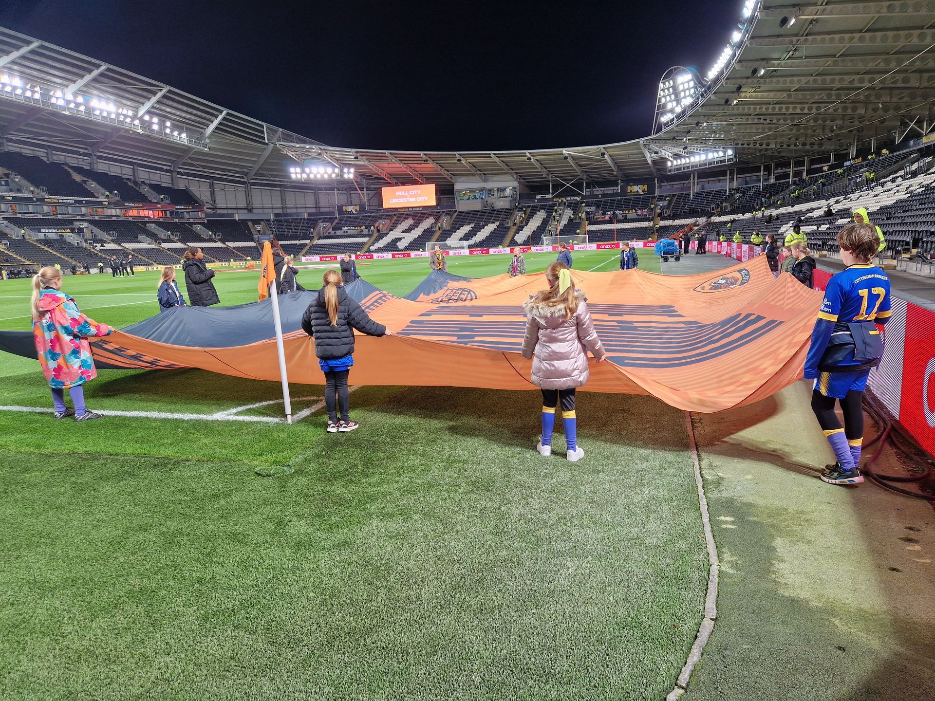 Children holding a large orange banner on a soccer field at night. Stadium in background.