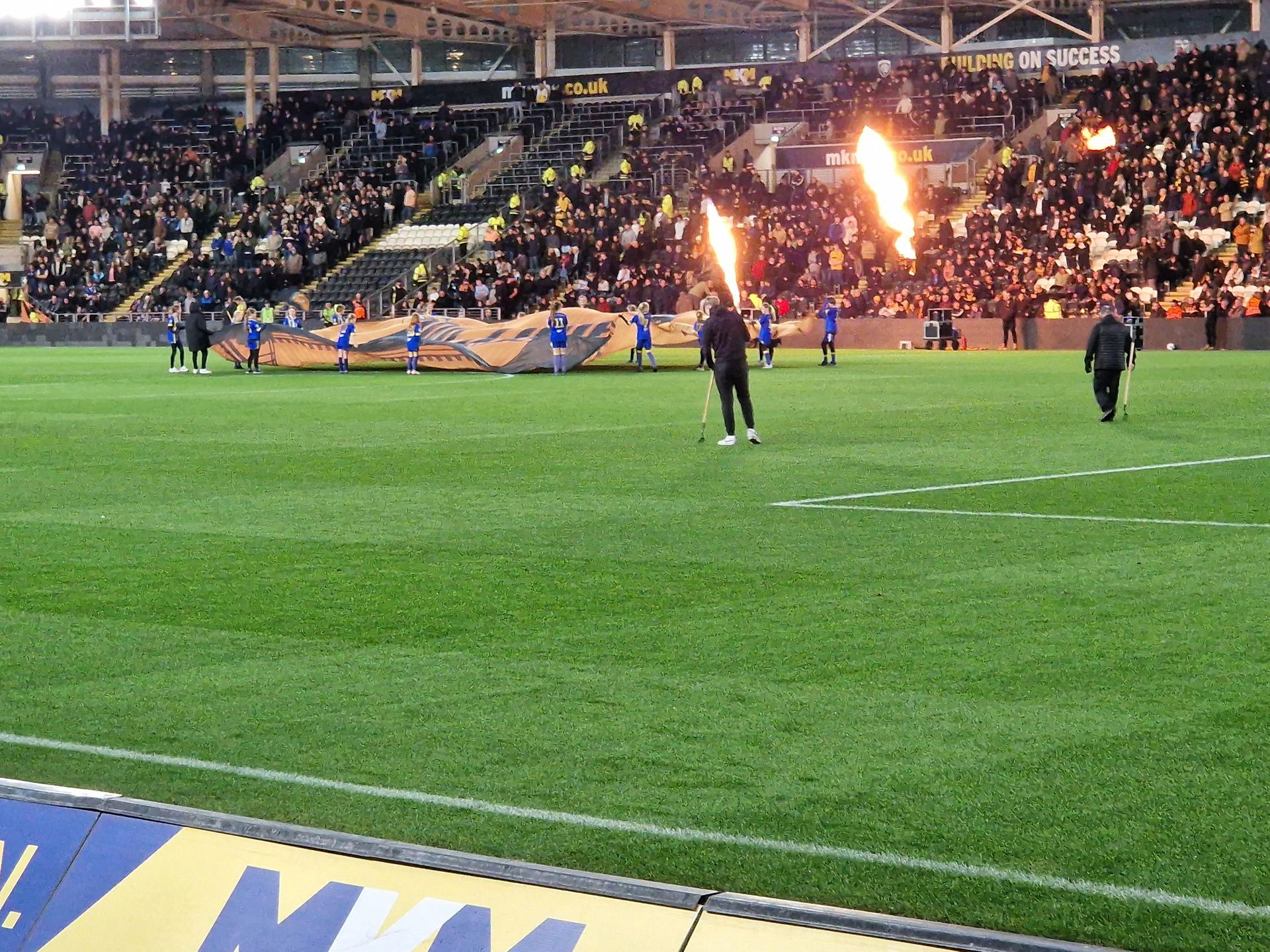 Soccer players on the field with fans in the stands, pyrotechnics erupt. Green field, black and gold seats.