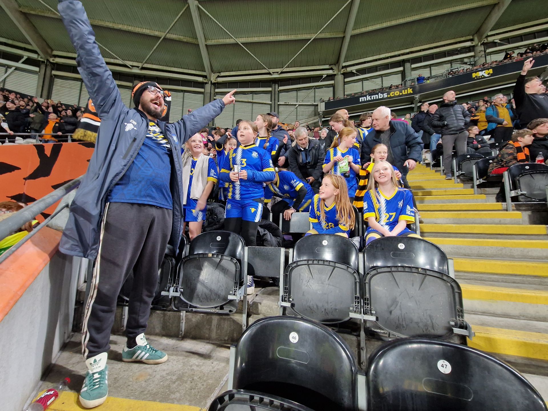 Fans cheer in a stadium. One man raises his arms, other fans wear blue jerseys, some empty seats.