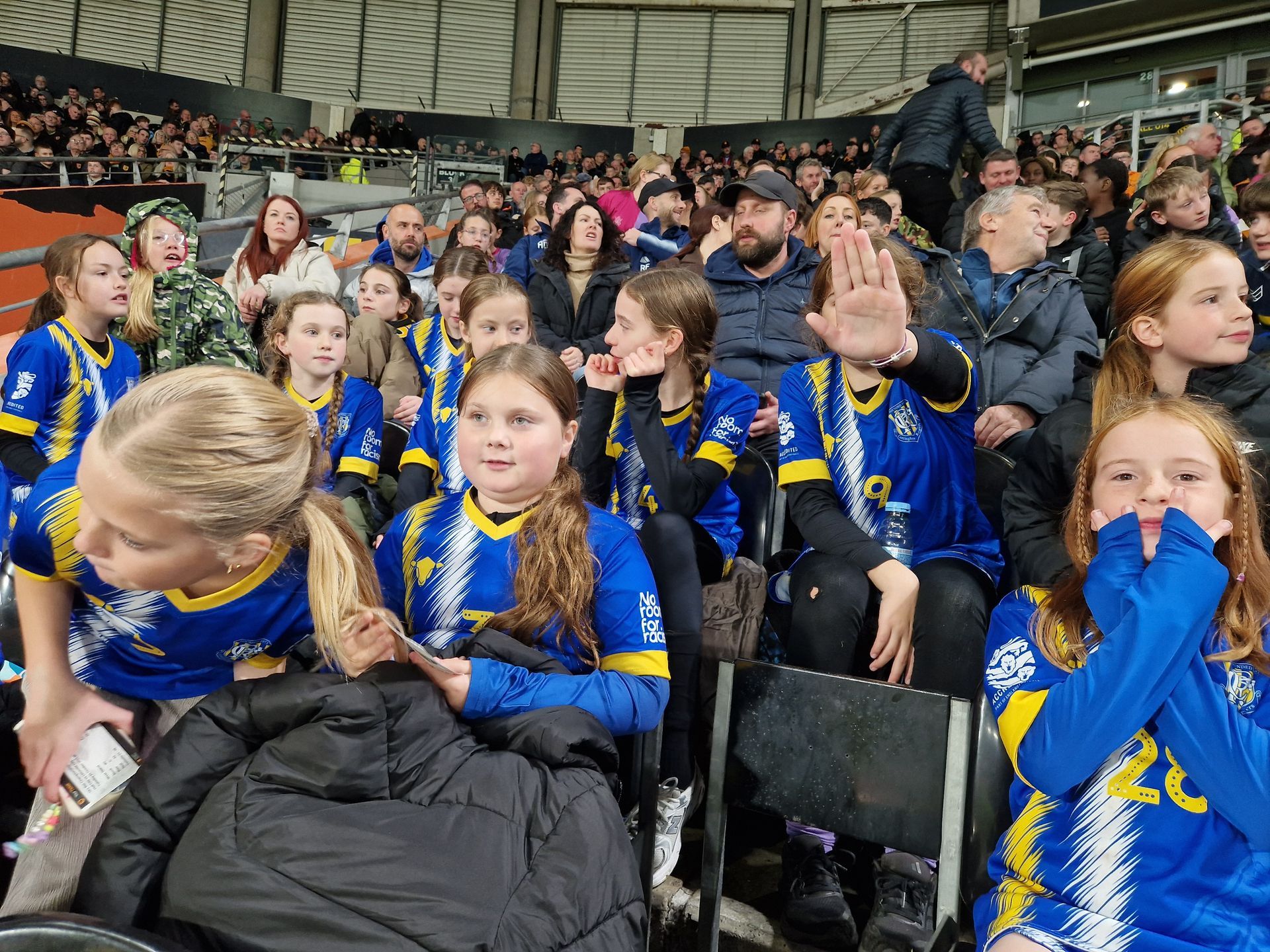 Group of children in blue and gold jerseys at an event, waving and smiling in a crowded indoor setting.
