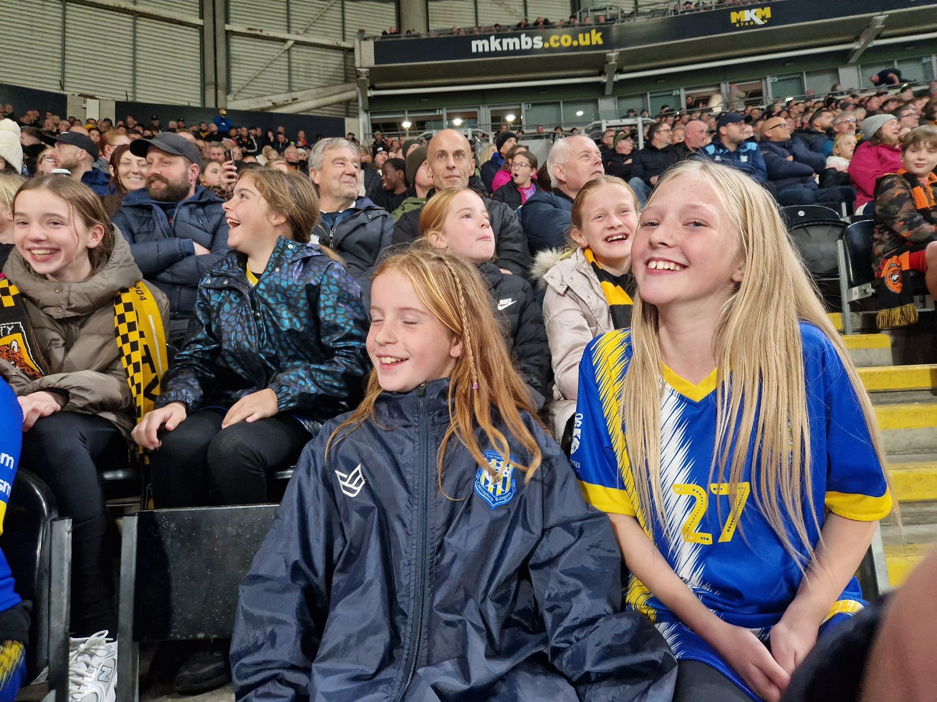 People in stadium seats, cheering. Two girls in blue and yellow shirts smile. Others watch the game.
