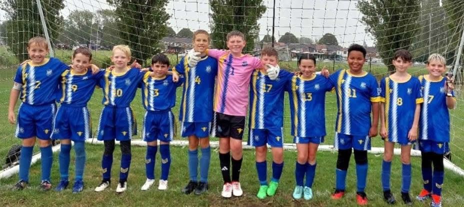 A youth soccer team in blue and white jerseys, lined up on a field, arms around each other, with a pink jersey in the middle.
