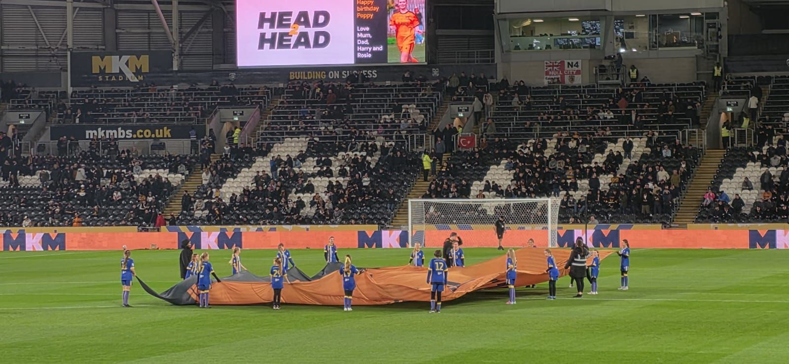 Soccer players on a field hold up a large sheet of orange tarp in front of a stadium crowd.
