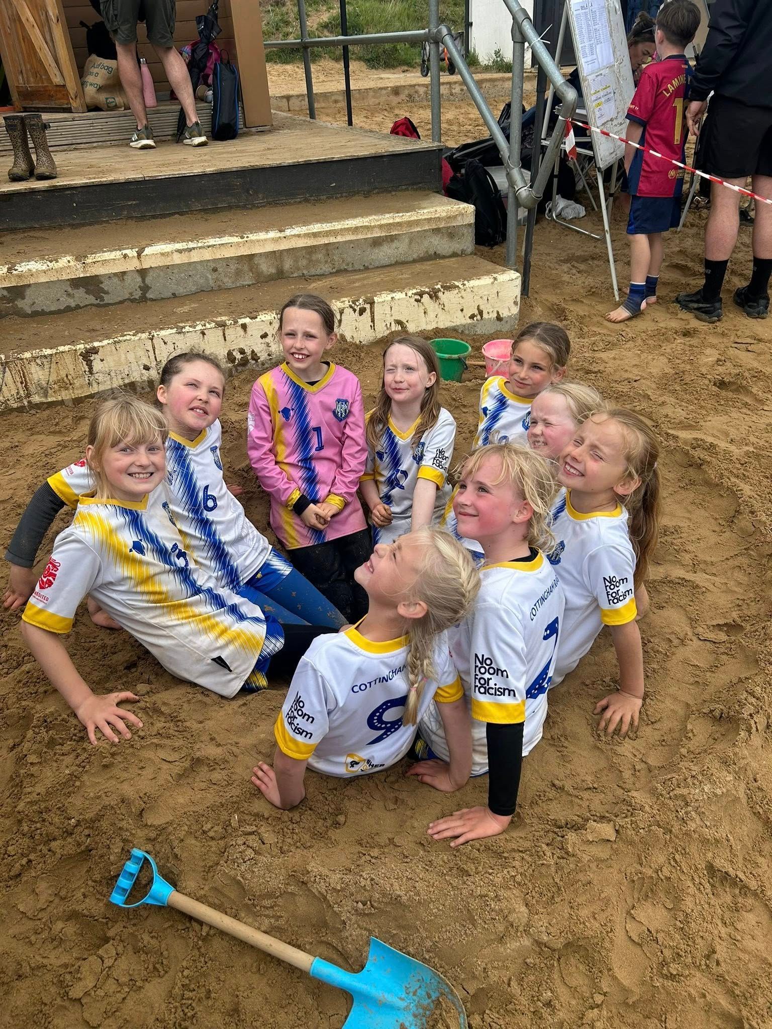 Girls in soccer jerseys in a sand pit, smiling. A shovel is in front.