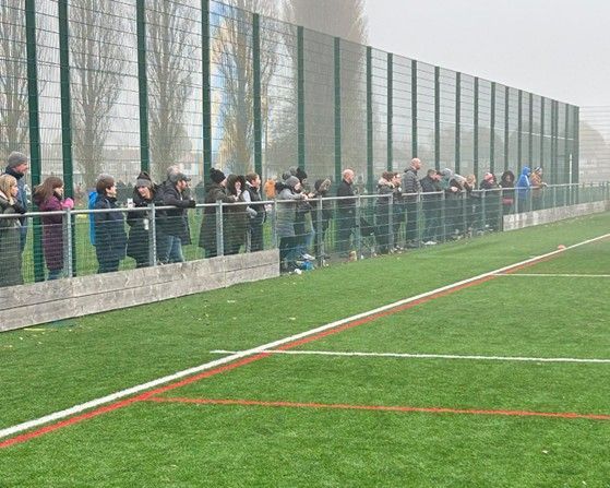 People watching a sporting event from behind a fence on a green field. Overcast day.