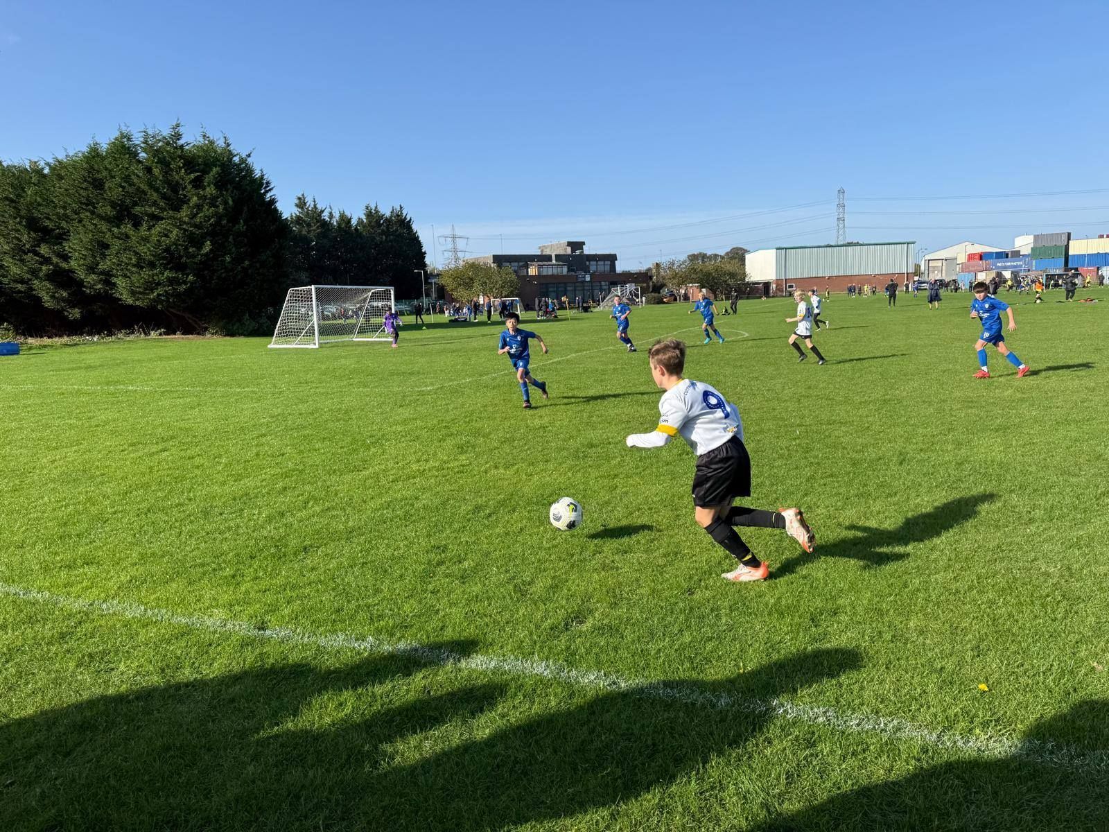 Soccer game on a green field under a clear blue sky. A player in a light blue jersey dribbles the ball.