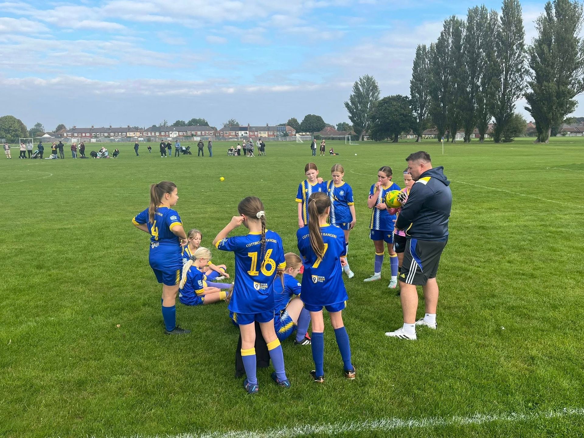 Girls' soccer team in blue and yellow jerseys huddle with a coach on a green field.