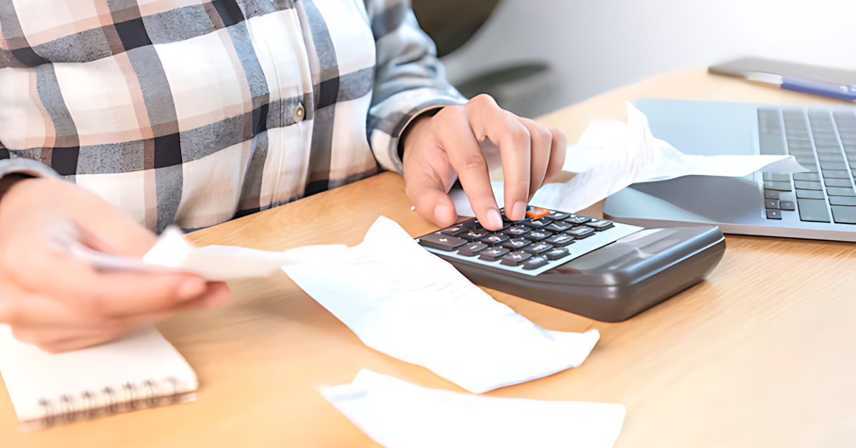 Person calculating bills with a calculator at a desk, with receipts and a laptop nearby