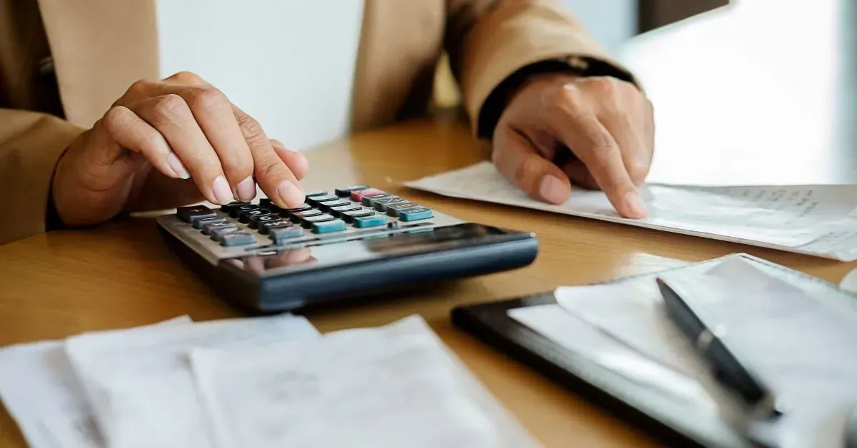 Hands using a calculator beside paperwork and a pen on a desk.