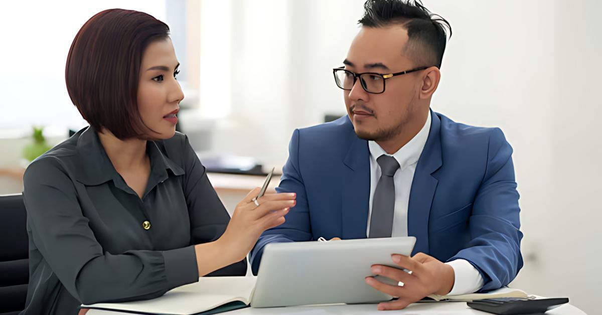 Two coworkers discuss a document at a desk, with a woman pointing at a tablet and a man listening attentively.