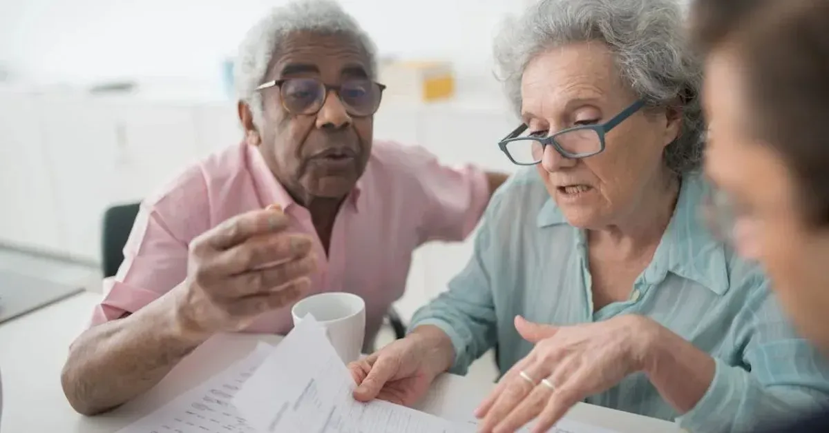 Two seniors reviewing papers together at a table, one holding a mug and pointing at documents.