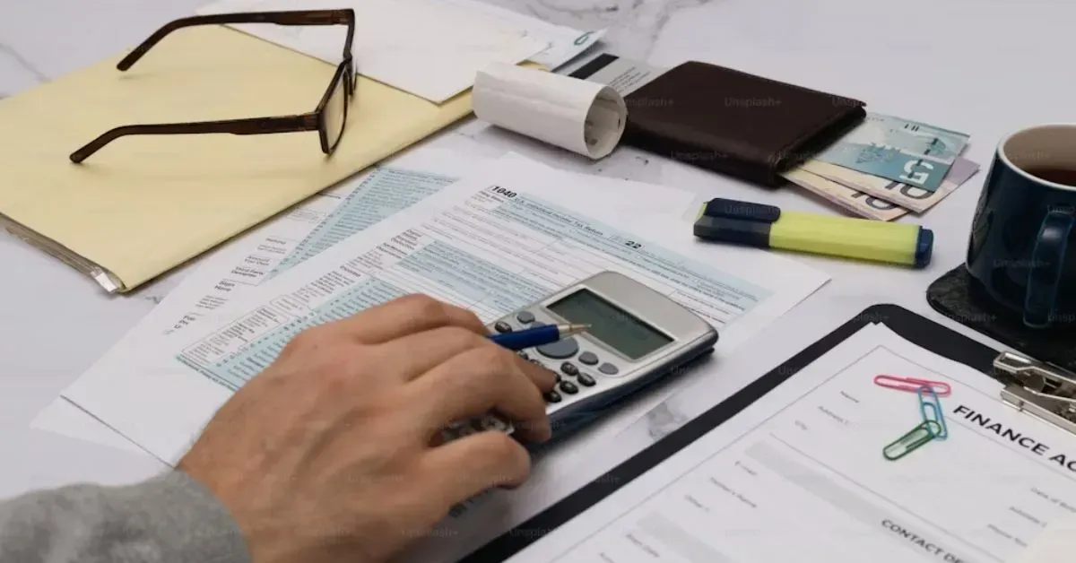 Hand using a calculator at a desk with paperwork, glasses, and office supplies