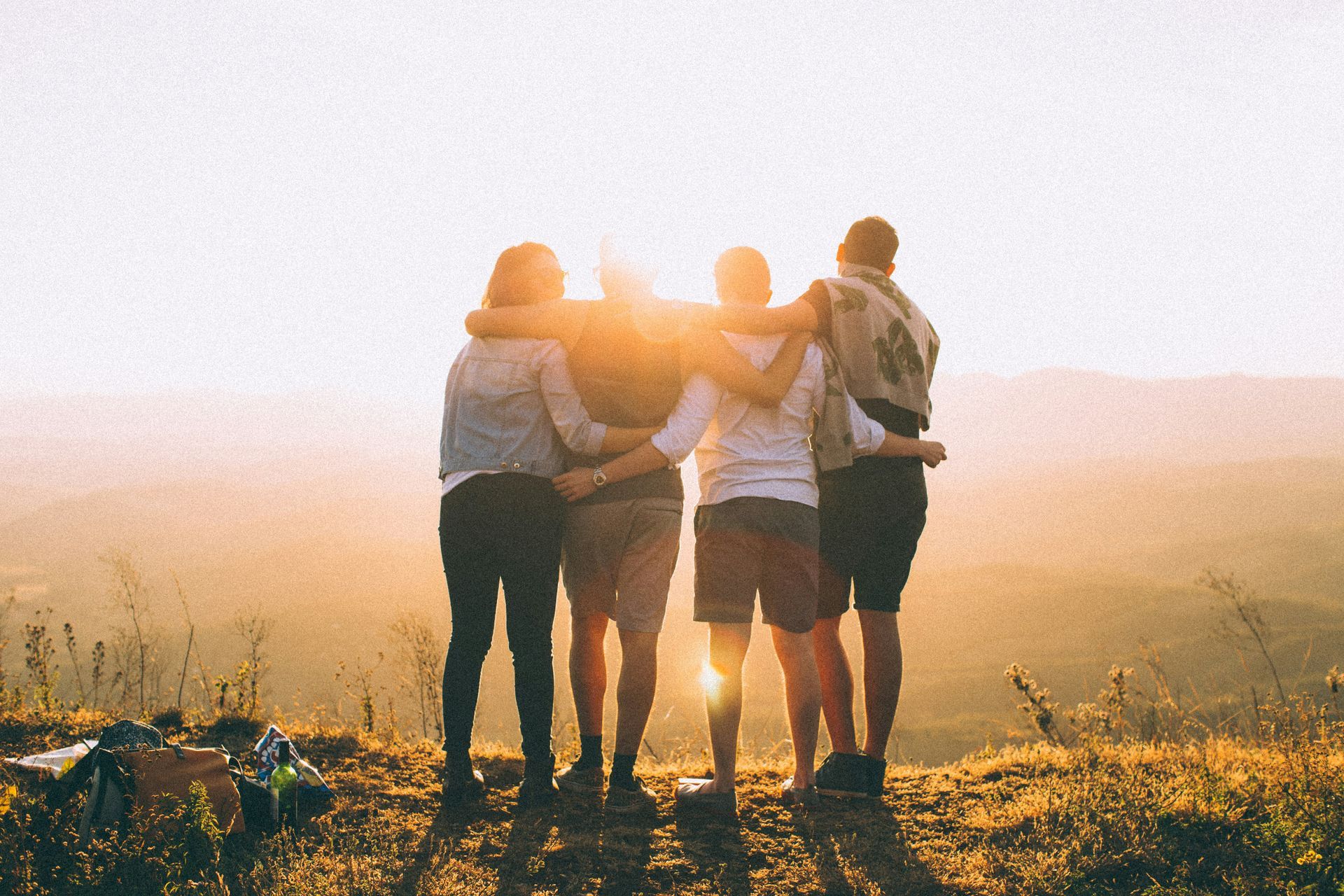 Four friends stand arm in arm on a hill at sunrise, facing a bright horizon.