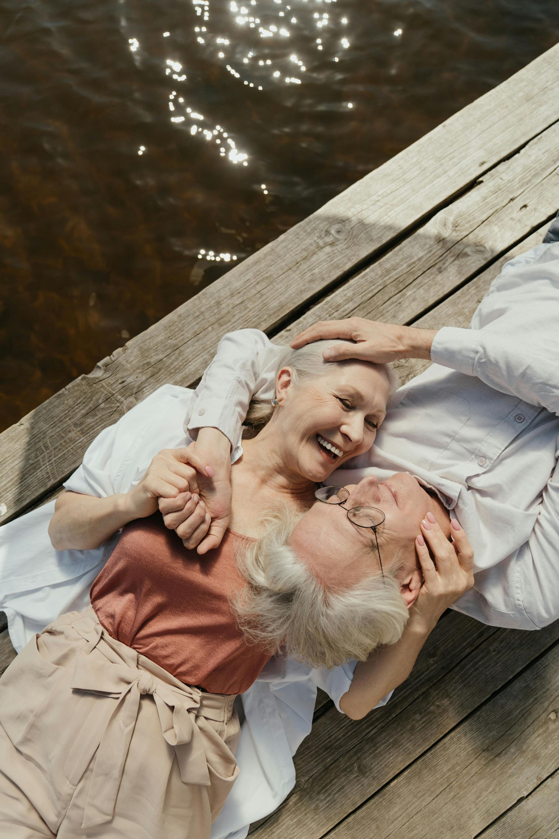 Two people lying on a wooden dock, smiling and holding hands beside dark water.
